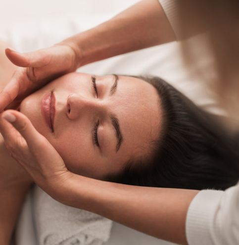 High angle of young female lying on bed with closed eyes while therapist makes a relaxing face massage procedure at spa